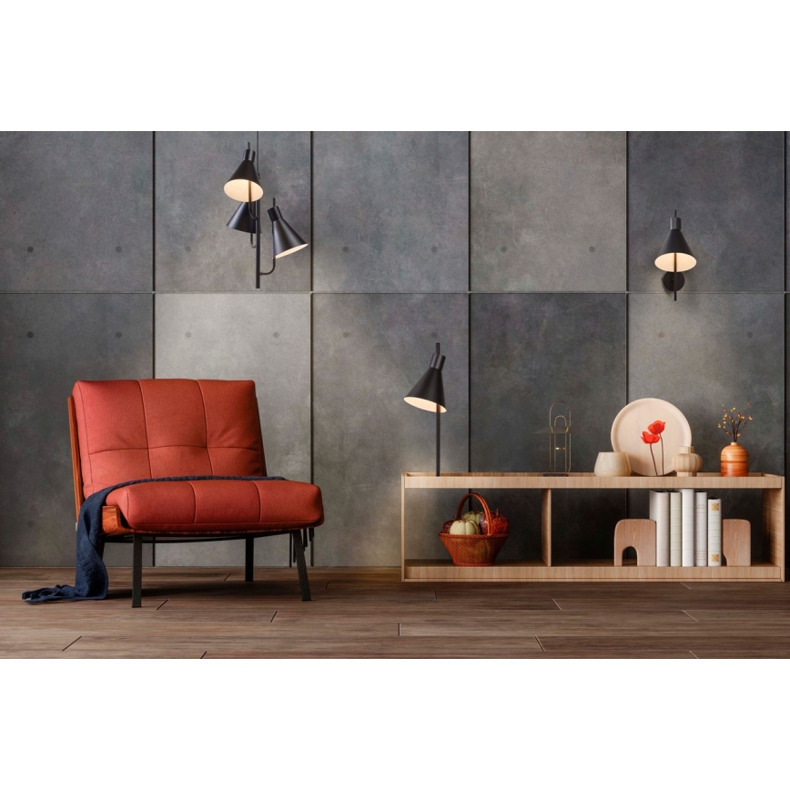 Červené kožené kreslo stojí v interiéri s drevenou podlahou. Na stene sú tri čierne nástenné lampy s kužeľovitými tienidlami. Vedľa kresla je drevená polica s dekoratívnymi predmetmi a kôšikom s ovocím.