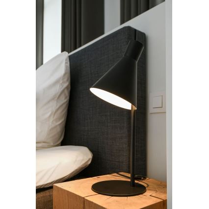 Stolová lampa s kuželovitým tienidlom svieti. Lampa je umiestnená na drevenom nočnom stolíku pri posteli.