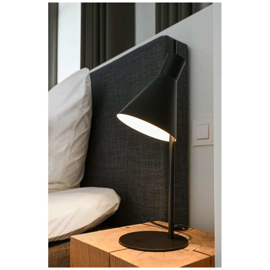 Stolová lampa s kuželovitým tienidlom svieti. Lampa je umiestnená na drevenom nočnom stolíku pri posteli.