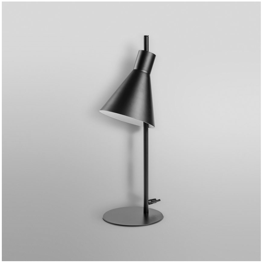 Stolová lampa s kužeľovitým tienidlom stojí na svetlom podklade. Lampa je čierna, s kovovou základňou. Tienidlo je mierne naklonené.