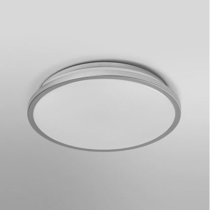 Okrúhle stropné svetlo je pripevnené k stropu. Svietidlo má biely difúzor a strieborný kovový rám. Svetlo je osadené na svetlom sivom strope.