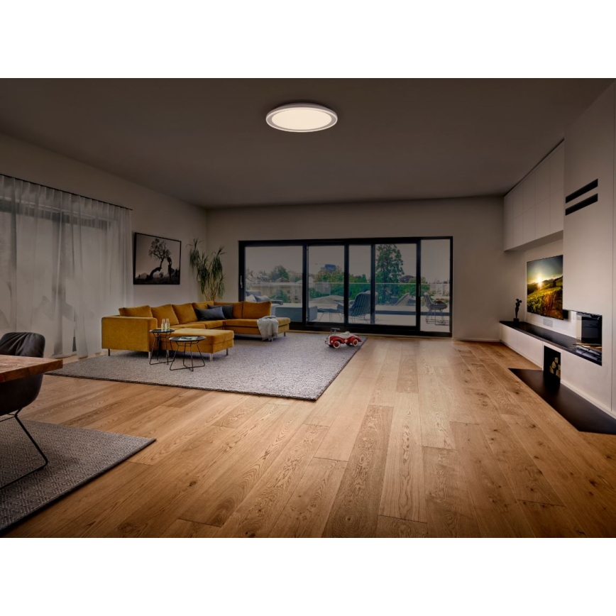 Svetlo svieti v priestrannej obývacej izbe s drevenou podlahou, žltou sedačkou a veľkými posuvnými dverami s výhľadom na exteriér. Na podlahe je rozložený koberec. V pozadí je viditeľná televízia a krb.