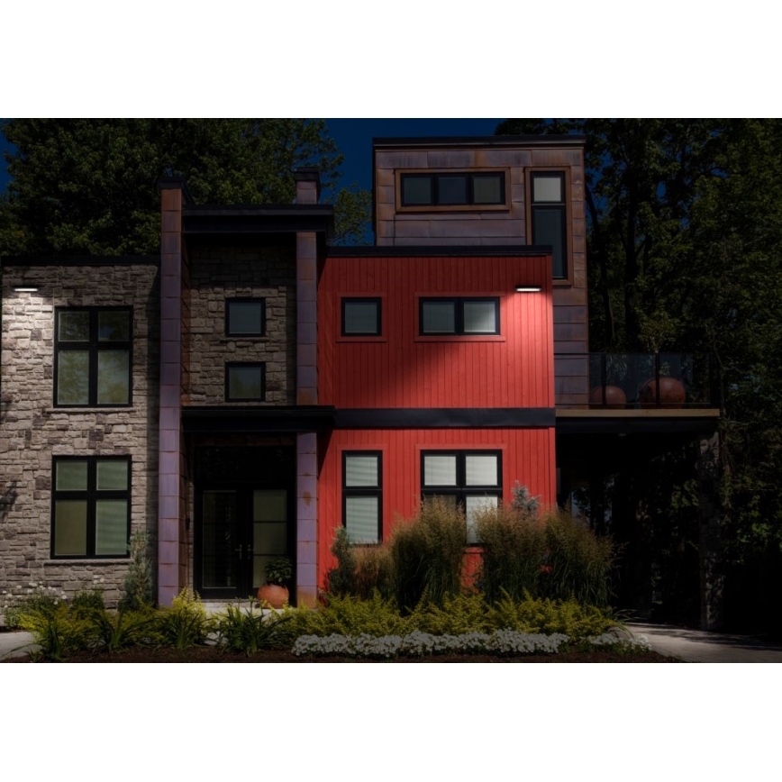 Vonkajšie nástenné svietidlo osvetľuje červenú fasádu moderného domu v noci. Dom je súčasťou radu domov s rôznymi farbami fasád a zeleňou pred ním.