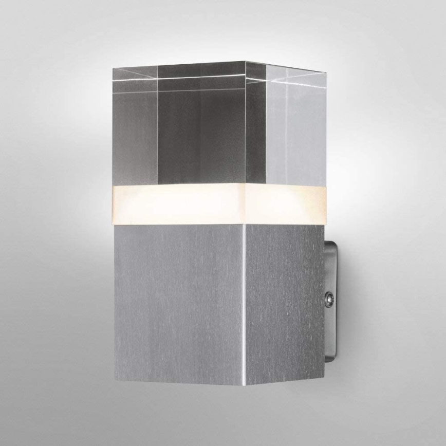 Nástenné svietidlo svieti. Svietidlo je kubické, pozostáva z kovového a skleneného prvku. Je umiestnené na svetlej stene.