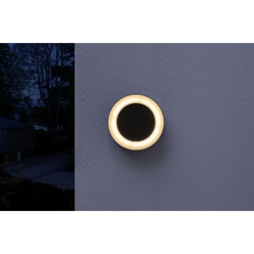 Okrúhle nástenné svietidlo svieti v noci. Svietidlo je pripevnené na sivú stenu. V pozadí je tmavá krajina.