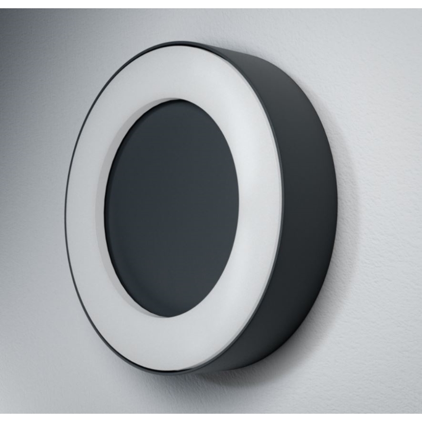 Okrúhly displej je pripevnený na stene. Má tmavosivý stred obklopený bielym kruhom a vonkajším tmavosivým okrajom.