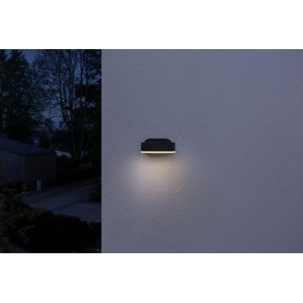 Čierna nástenná lampa svieti na svetlej stene. Lampa je v tvare obdĺžnika. Pozadie je tmavá noc.