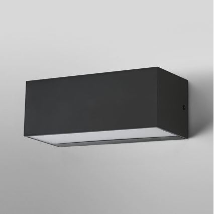 Obdĺžniková nástenná lampa svieti. Je tmavosivá, pripevnená k stene. Spodná časť lampy je jemne presvetlená.