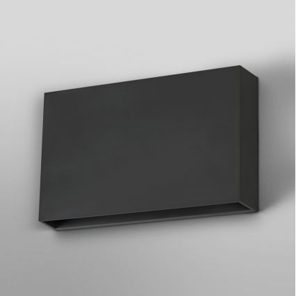 Obdĺžniková nástenná lampa s tmavosivou povrchovou úpravou je pripevnená na stene. Svietidlo má v spodnej časti úzky otvor. Svetlošedá stena tvorí pozadie.