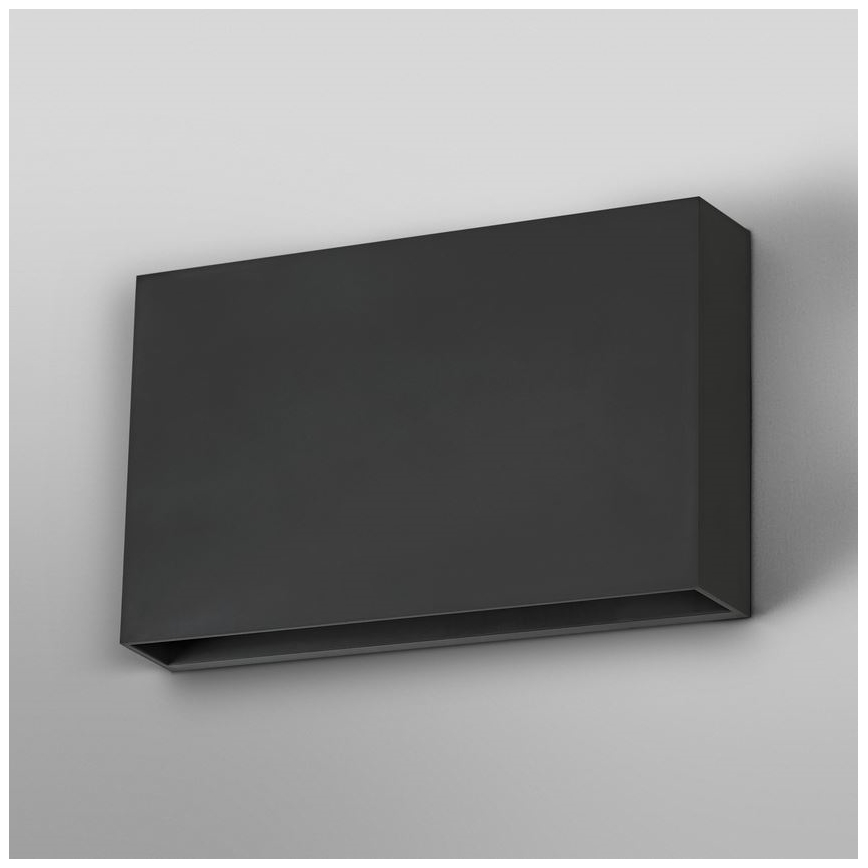 Obdĺžniková nástenná lampa s tmavosivou povrchovou úpravou je pripevnená na stene. Svietidlo má v spodnej časti úzky otvor. Svetlošedá stena tvorí pozadie.