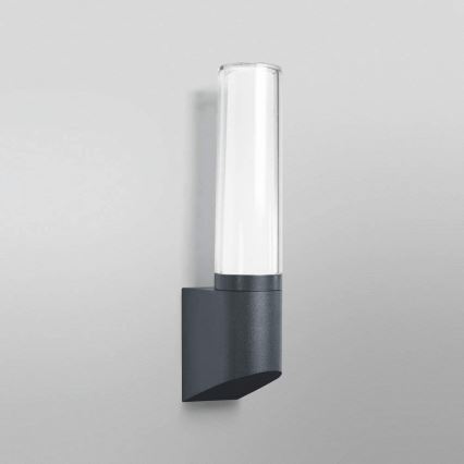 Svietidlo svieti na stene. Je to vertikálne umiestnené nástenné svietidlo s valcovitým skleneným difúzorom a tmavosivou kovovou základňou.