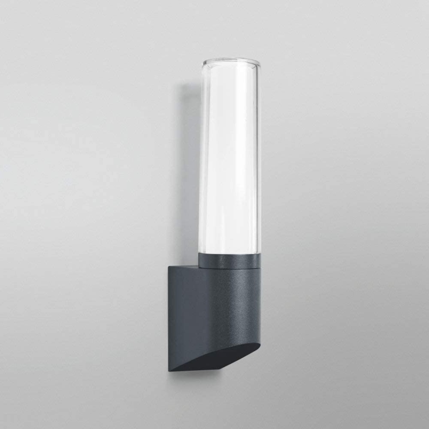 Svietidlo svieti na stene. Je to vertikálne umiestnené nástenné svietidlo s valcovitým skleneným difúzorom a tmavosivou kovovou základňou.
