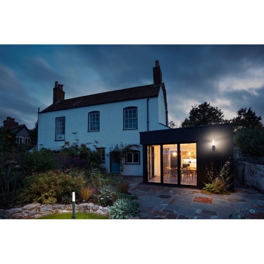 Vonkajší nástenný svietidlo svieti na zadnej strane domu s prístavbou. Dom je biely, prístavba tmavá. Okolité prostredie tvorí osvetlená záhrada v súmraku.