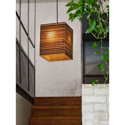 Kubická drevená lampa visí zo stropu. Svieti v interiéri s dreveným schodiskom a bielou tehlovou stenou. V pozadí je zelená rastlina.