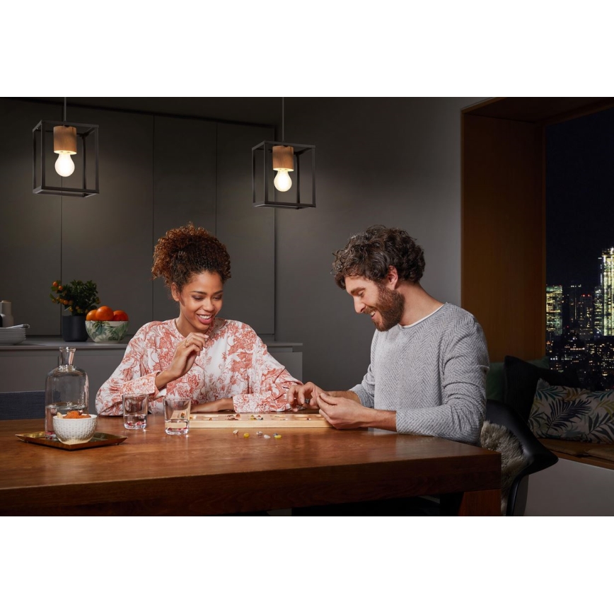 Pár hrá spoločenskú hru pri drevenom stole osvetlenom dvoma závesnými lampami v modernom interiéri s výhľadom na nočné mesto.