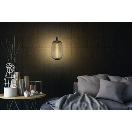 Závesná lampa s tmavým skleneným tienidlom v tvare kapsuly svieti v interiéri. Lampa visí nad nízkym stolíkom s dekoráciami a vedľa pohovky s vankúšmi a dekou.