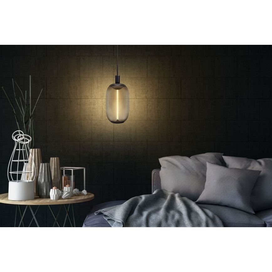 Závesná lampa s tmavým skleneným tienidlom v tvare kapsuly svieti v interiéri. Lampa visí nad nízkym stolíkom s dekoráciami a vedľa pohovky s vankúšmi a dekou.