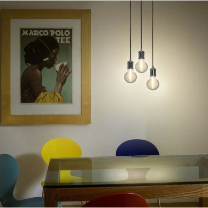 Tri závesné žiarovky svietia nad jedálenským stolom. Stolík je sklenený, obklopený farebnými stoličkami. Na stene visí obraz s nápisom „MARCO POLO TEE“.