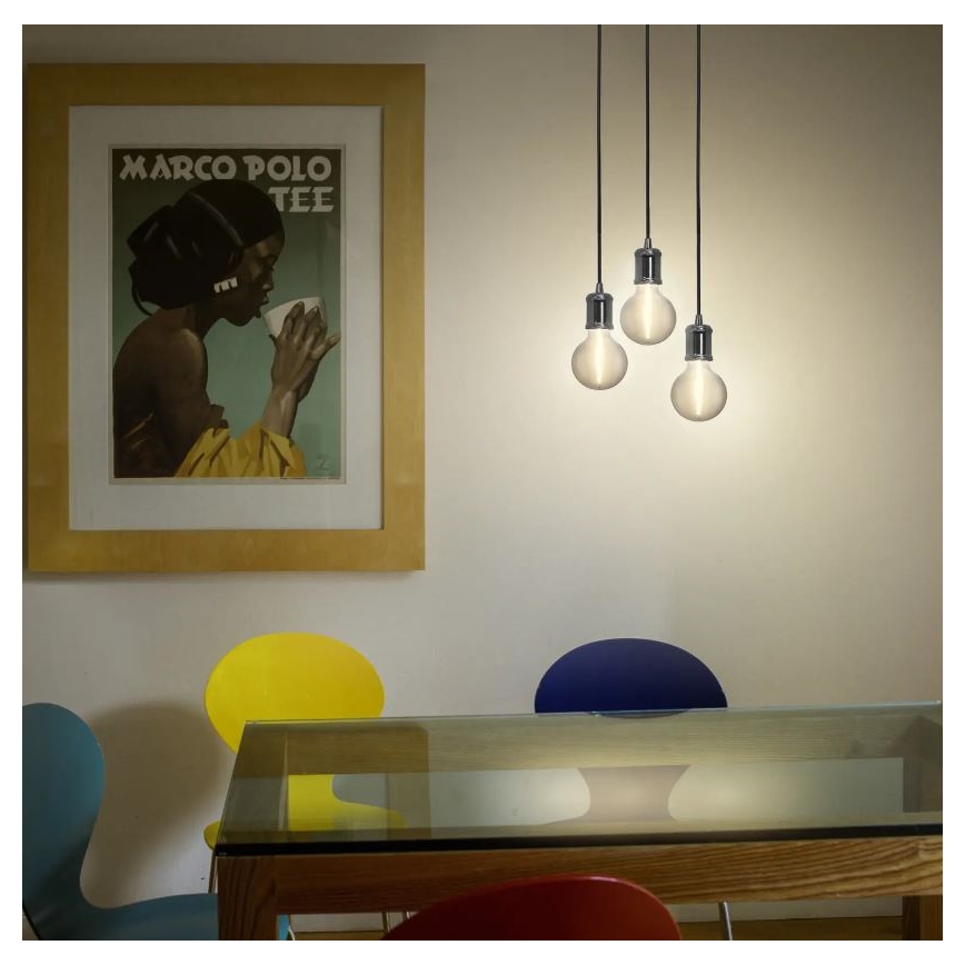 Tri závesné žiarovky svietia nad jedálenským stolom. Stolík je sklenený, obklopený farebnými stoličkami. Na stene visí obraz s nápisom „MARCO POLO TEE“.