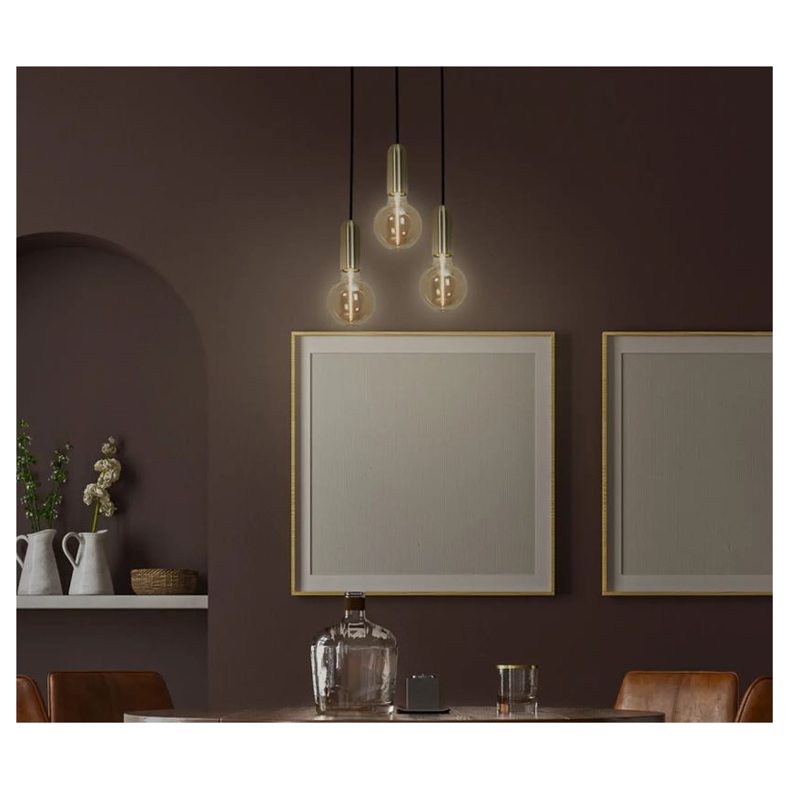 Trojramenné závesné svietidlo s guľovitými tienidlami svieti v tmavej miestnosti. Svietidlo je umiestnené nad stolom s dekoráciami. V pozadí sú obrazy v zlatých rámoch.