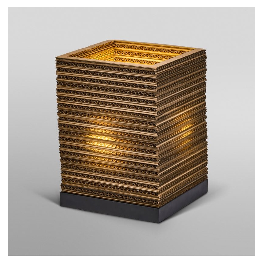 Stolová lampa z prekladaných vrstiev kartónu svieti. Lampa má štvorcový tvar a stojí na tmavej podstavci. Svetlo presvitá cez vrstvy.