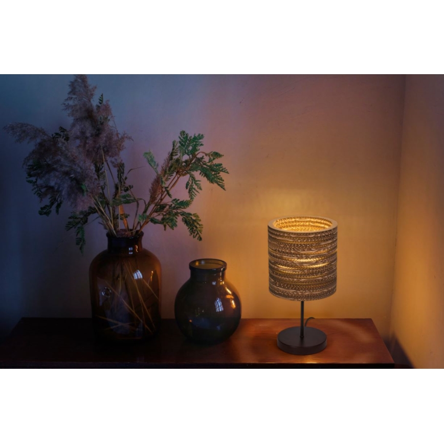 Stolová lampa s valcovým tienidlom z recyklovaného materiálu svieti na tmavom drevenom povrchu. Vedľa nej stoja dve vázy s kvetmi.