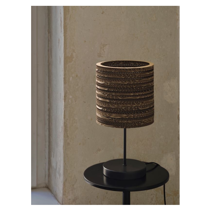 Stolová lampa s valcovitým tienidlom z vlnitého kartónu stojí na čiernom stolíku. Tienidlo je zložené z vodorovne prepletených pásikov kartónu. Lampa má čierny kovový stojan a základňu.