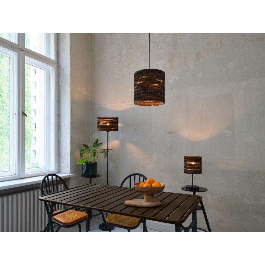 Svietidlá z kartónu svietia v jedálni. Závesné svietidlo je umiestnené nad stolom, stojacia lampa a stolová lampa sú vedľa stola. V pozadí je okno a betónová stena.