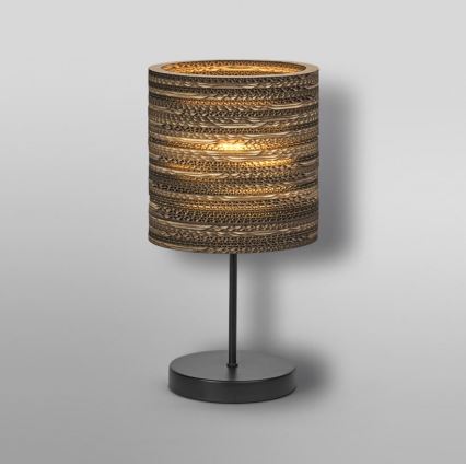 Stolová lampa svieti. Tienidlo je valcovité, vyrobené z vlnitého kartónu, hnedo-zlatej farby. Podstavec je čierny a kovový. Lampa je umiestnená na svetlom pozadí.