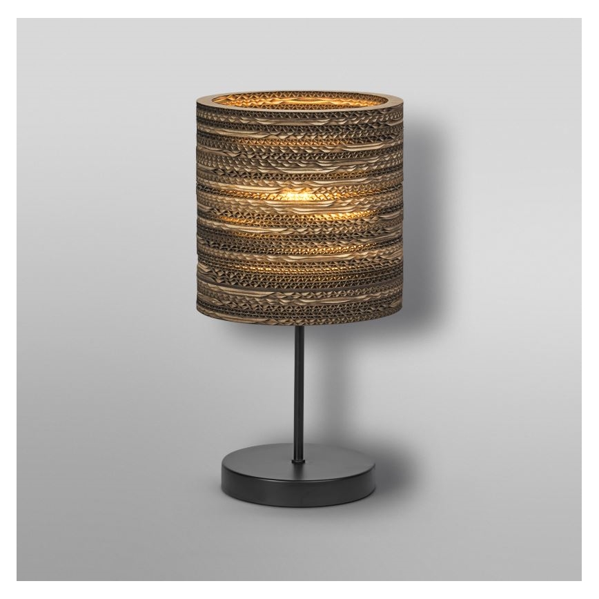 Stolová lampa svieti. Tienidlo je valcovité, vyrobené z vlnitého kartónu, hnedo-zlatej farby. Podstavec je čierny a kovový. Lampa je umiestnená na svetlom pozadí.