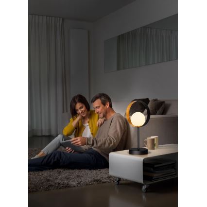 Stolová lampa s kruhovým tienidlom svieti v obývacej izbe. Pár sedí na koberci a pozerá sa na tablet. Lampa stojí na nízkom bielom stolíku.