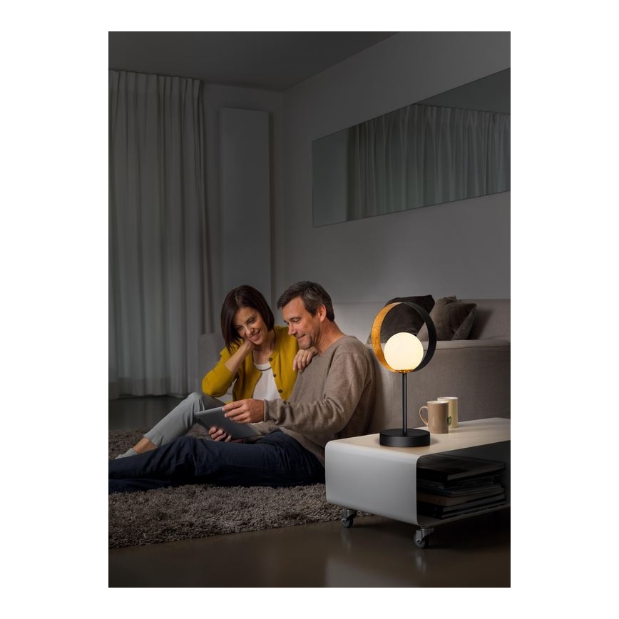 Stolová lampa s kruhovým tienidlom svieti v obývacej izbe. Pár sedí na koberci a pozerá sa na tablet. Lampa stojí na nízkom bielom stolíku.