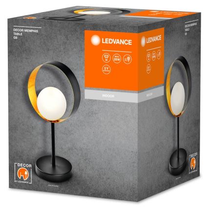 Stolová lampa s čiernym kovový rámom a zlatou povrchovou úpravou, má bielu guľovú tienidlo. Nachádza sa v kartónovej krabici s nápismi „LEDVANCE“, „DECOR MEMPHIS TABLE G9“, „MAX 28W“, „INDOOR“ a „5Y“.
