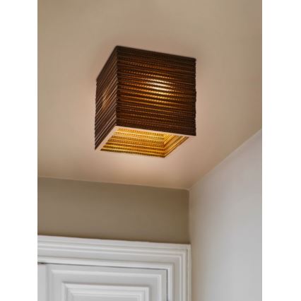 Kubická lampa zo svetlohnedého materiálu s vnútorným zlatým rámom svieti pripevnená k stropu v rohu miestnosti pri dverách.