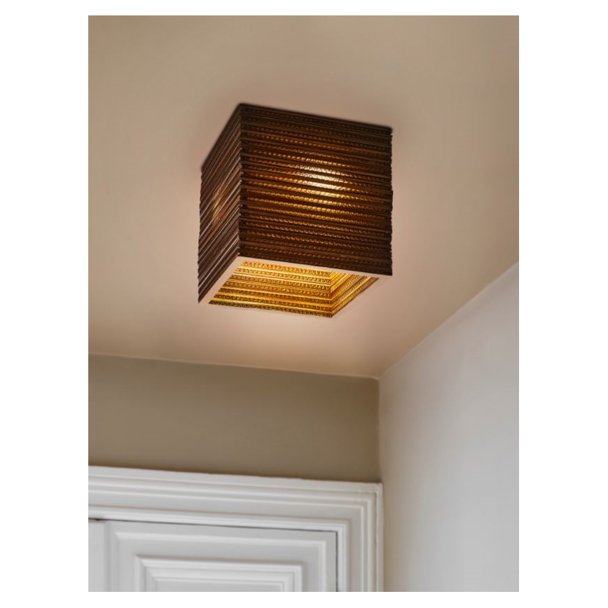 Kubická lampa zo svetlohnedého materiálu s vnútorným zlatým rámom svieti pripevnená k stropu v rohu miestnosti pri dverách.