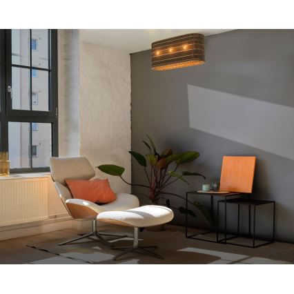 Obdĺžnikové stropné svietidlo svieti v interiéri. Svietidlo je pletené, s tromi svetlami. V miestnosti je kreslo s podnožkou a stolík.