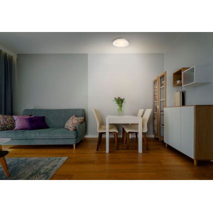 Svietidlo svieti v obývacej izbe. Nachádza sa na strope nad jedálenským stolom a pohovkou. Izba má drevenú podlahu, sivé steny a nábytok v svetlých farbách.