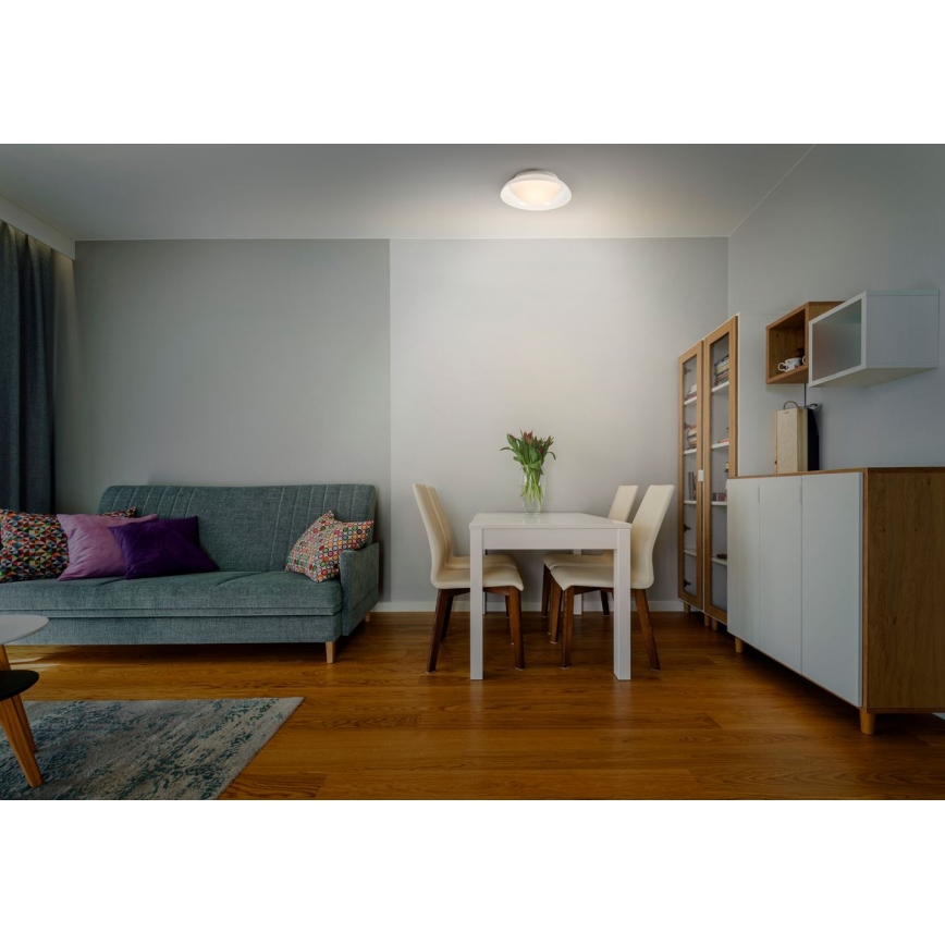 Svietidlo svieti v obývacej izbe. Nachádza sa na strope nad jedálenským stolom a pohovkou. Izba má drevenú podlahu, sivé steny a nábytok v svetlých farbách.