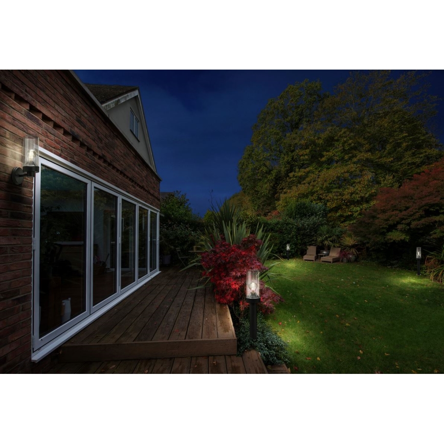 Záhradné osvetlenie svieti v nočnom prostredí. Svietidlá sú umiestnené na stene domu, na terase a v trávniku. Dom má drevenú terasu a veľké francúzske okná.