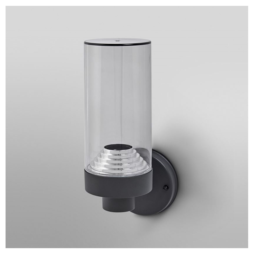 Nástenná lampa svieti. Sklenený valec chráni kovový odrazový prvok. Upevnenie je tmavosivé, na svetlej stene.