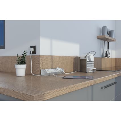 Predlžovací kábel s tromi zásuvkami je zapojený do elektrickej zásuvky v kuchynskej linke. K predlžovačke sú pripojené káble nabíjačky telefónu a kávovaru. Kávovar a telefón ležia na drevenom kuchynskom stole.