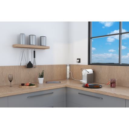 Trojpólový predlžovací kábel je zapojený do zásuvky v kuchynskej linke. Kábel napája kávovar, ktorý stojí na kuchynskej linke blízko okna s výhľadom na oblohu.