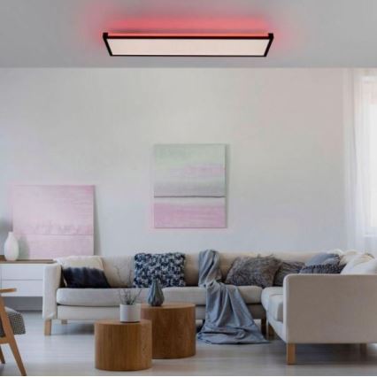 Stropné svietidlo svieti v obývacej izbe. Svietidlo je obdĺžnikové, s červeným podsvietením. Izba obsahuje rohovú pohovku, konferenčné stolíky a obrazy na stenách.