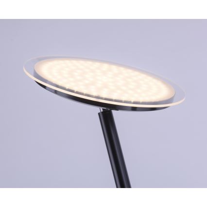 Stojacia lampa svieti. Plochá, okrúhla LED hlavica svietidla je umiestnená na čiernej kovovej nohe. Svetlo je rozptýlené cez priesvitný kryt.