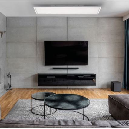 Televízor je umiestnený na čiernej televíznej skrínke pred betónovou stenou v obývacej izbe. Pod televízorom je soundbar. V miestnosti sú dva konferenčné stolíky a koberec.