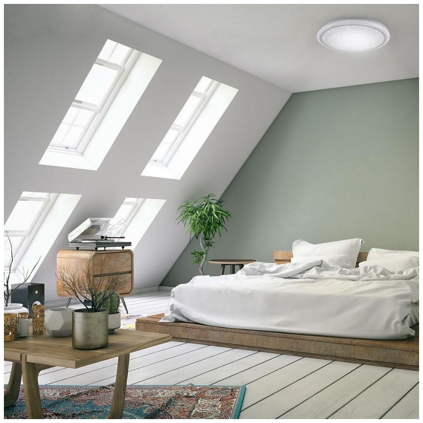 Moderná spálňa s posteľou a drevenou platformou, osvetlená stropným svietidlom. V miestnosti sú šikmé steny so strešnými oknami a nábytok v minimalistickom štýle.