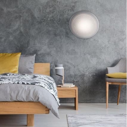 Drevená posteľ s posteľnou bielizňou je umiestnená v spálni. Na stene je pripevnené kruhové nástenné svietidlo. Vedľa postele stojí nočný stolík s lampou.