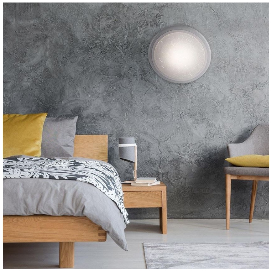 Drevená posteľ s posteľnou bielizňou je umiestnená v spálni. Na stene je pripevnené kruhové nástenné svietidlo. Vedľa postele stojí nočný stolík s lampou.