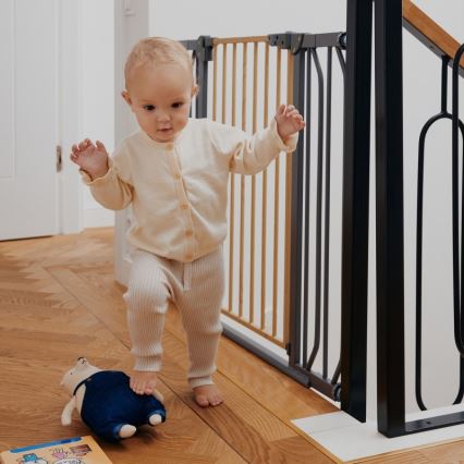 Dieťa v svetlom oblečení kráča po drevenej podlahe pri bezpečnostnej bránke na schodoch. Pri nohách má plyšovú hračku.
