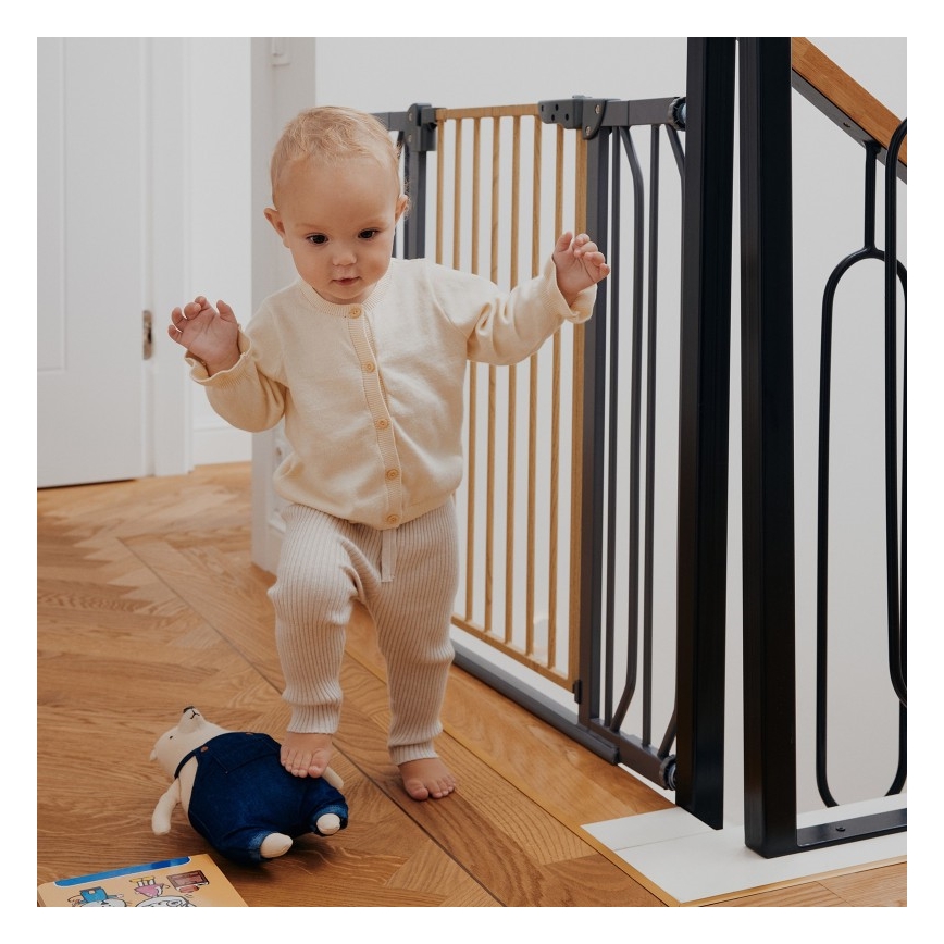 Dieťa v svetlom oblečení kráča po drevenej podlahe pri bezpečnostnej bránke na schodoch. Pri nohách má plyšovú hračku.